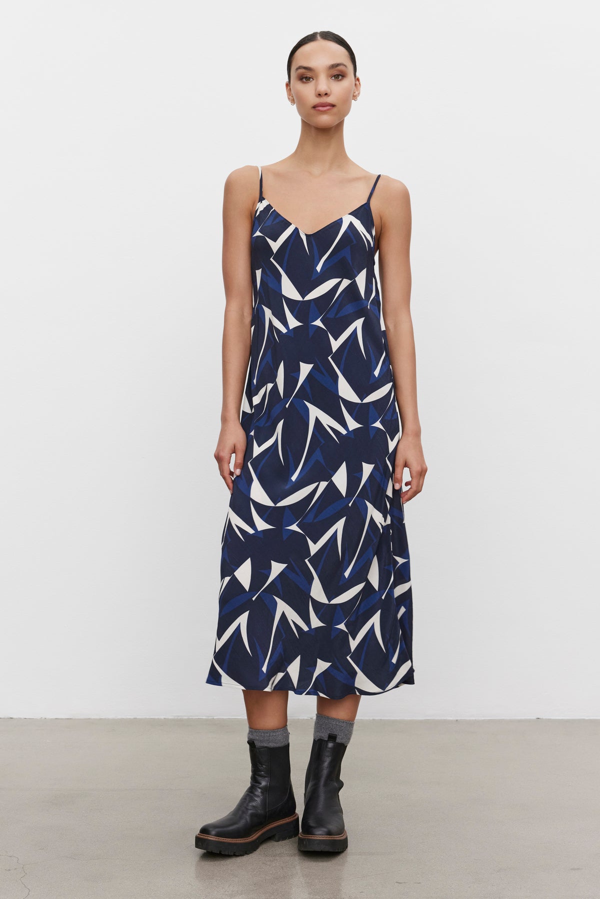   A person wearing the PERRY DRESS from Velvet by Graham & Spencer, characterized by a blue and white print with adjustable straps and crafted from royal crepe fabric, paired with black ankle boots, is standing against a plain white background. 