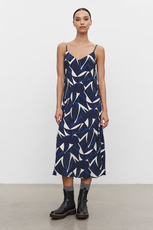 A person wearing the PERRY DRESS from Velvet by Graham & Spencer, characterized by a blue and white print with adjustable straps and crafted from royal crepe fabric, paired with black ankle boots, is standing against a plain white background.