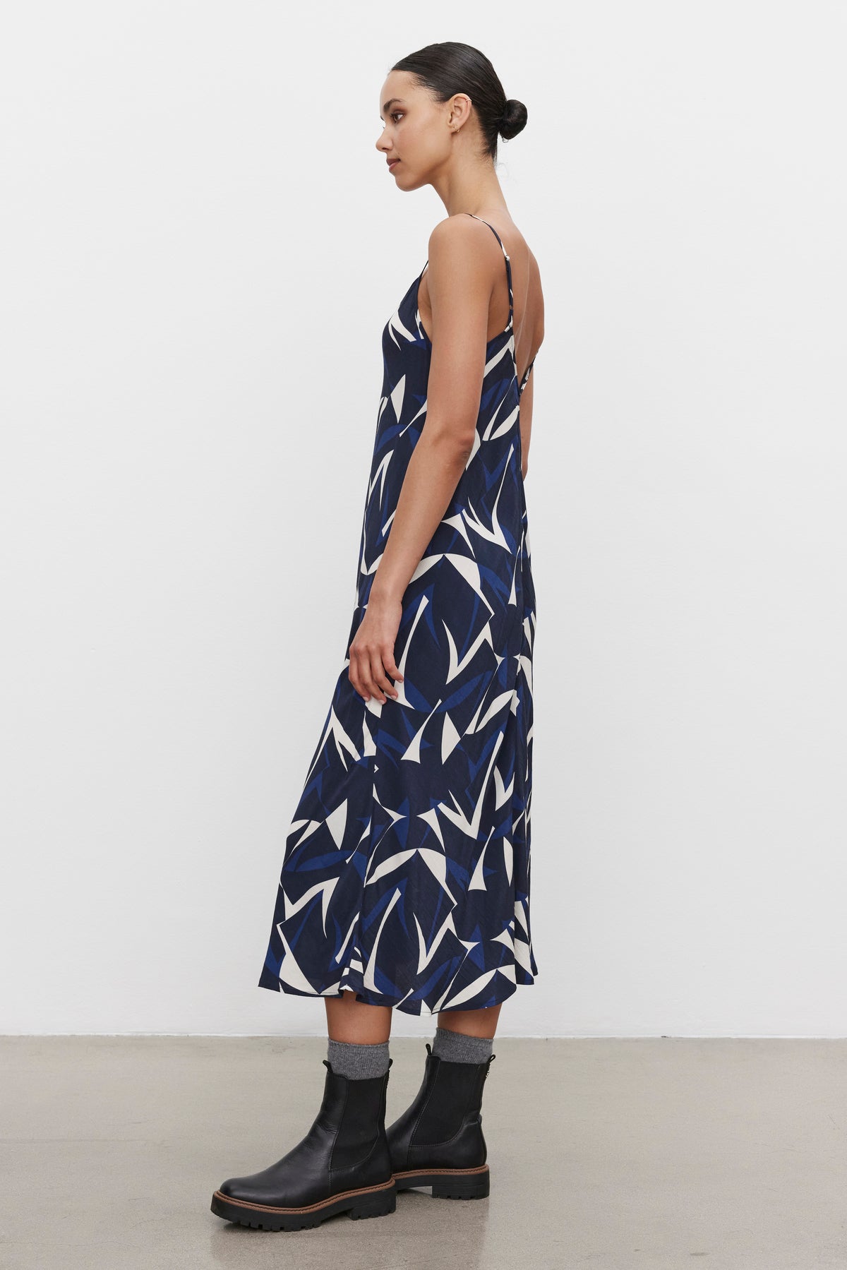   Person stands sideways, showcasing the PERRY DRESS from Velvet by Graham & Spencer, a sleeveless navy and white patterned tank dress with adjustable straps. The outfit is paired with black ankle boots and grey socks against a plain white background. 