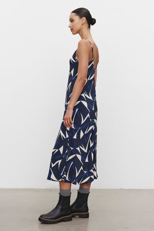 Person stands sideways, showcasing the PERRY DRESS from Velvet by Graham & Spencer, a sleeveless navy and white patterned tank dress with adjustable straps. The outfit is paired with black ankle boots and grey socks against a plain white background.