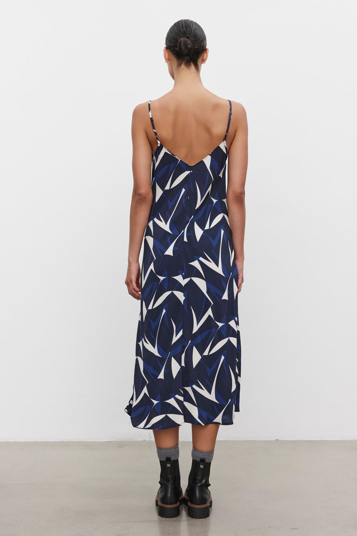   A person with dark hair in a low bun is standing with their back facing the camera. They are wearing the Velvet by Graham & Spencer PERRY DRESS, a sleeveless navy tank dress with a white abstract pattern, paired with black ankle boots. 