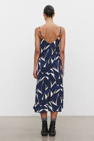 A person with dark hair in a low bun is standing with their back facing the camera. They are wearing the Velvet by Graham & Spencer PERRY DRESS, a sleeveless navy tank dress with a white abstract pattern, paired with black ankle boots.