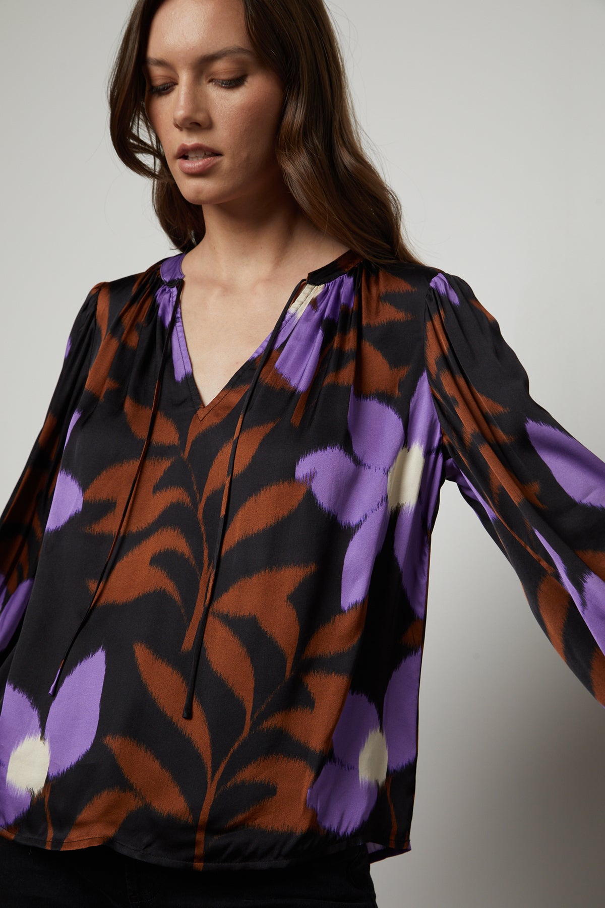 A woman wearing an ISRA PRINTED V-NECK BLOUSE by Velvet by Graham & Spencer with a modern print in purple and black floral.-35503650373825