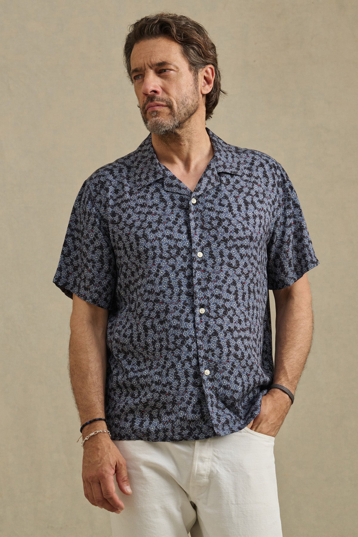 A man wears the EMILIO SHIRT by Velvet by Graham & Spencer— a short sleeve, breathable modal camp collar shirt—paired with white pants, standing with one hand in his pocket against a plain background.