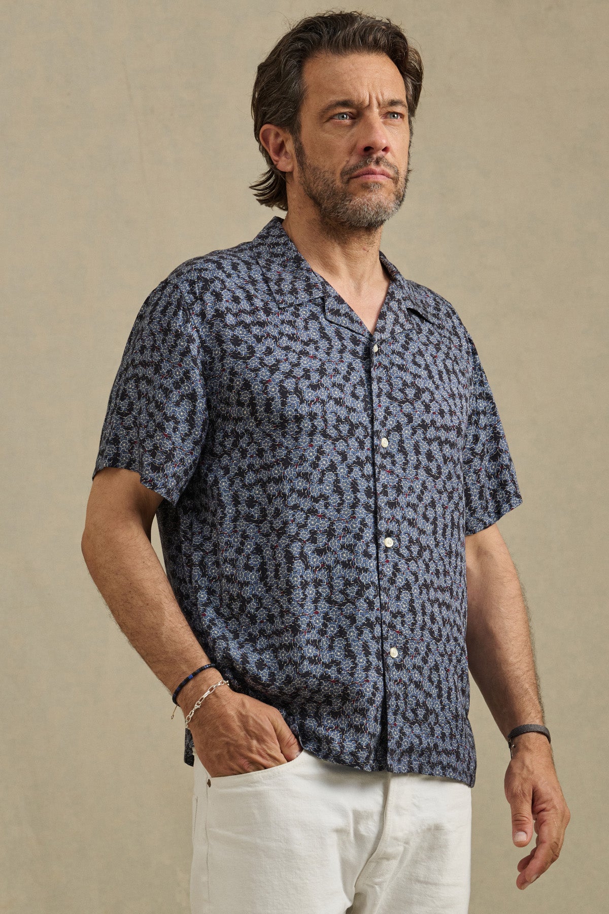 A man stands against a plain background wearing the EMILIO SHIRT by Velvet by Graham & Spencer—a breathable modal camp collar shirt with short sleeves—paired with white pants, one hand in his pocket.