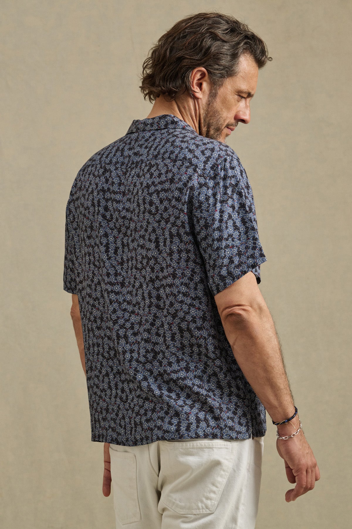A man with graying hair stands sideways against a plain beige background, wearing the EMILIO SHIRT by Velvet by Graham & Spencer—a patterned navy camp collar shirt made from breathable modal—paired with light-colored pants.