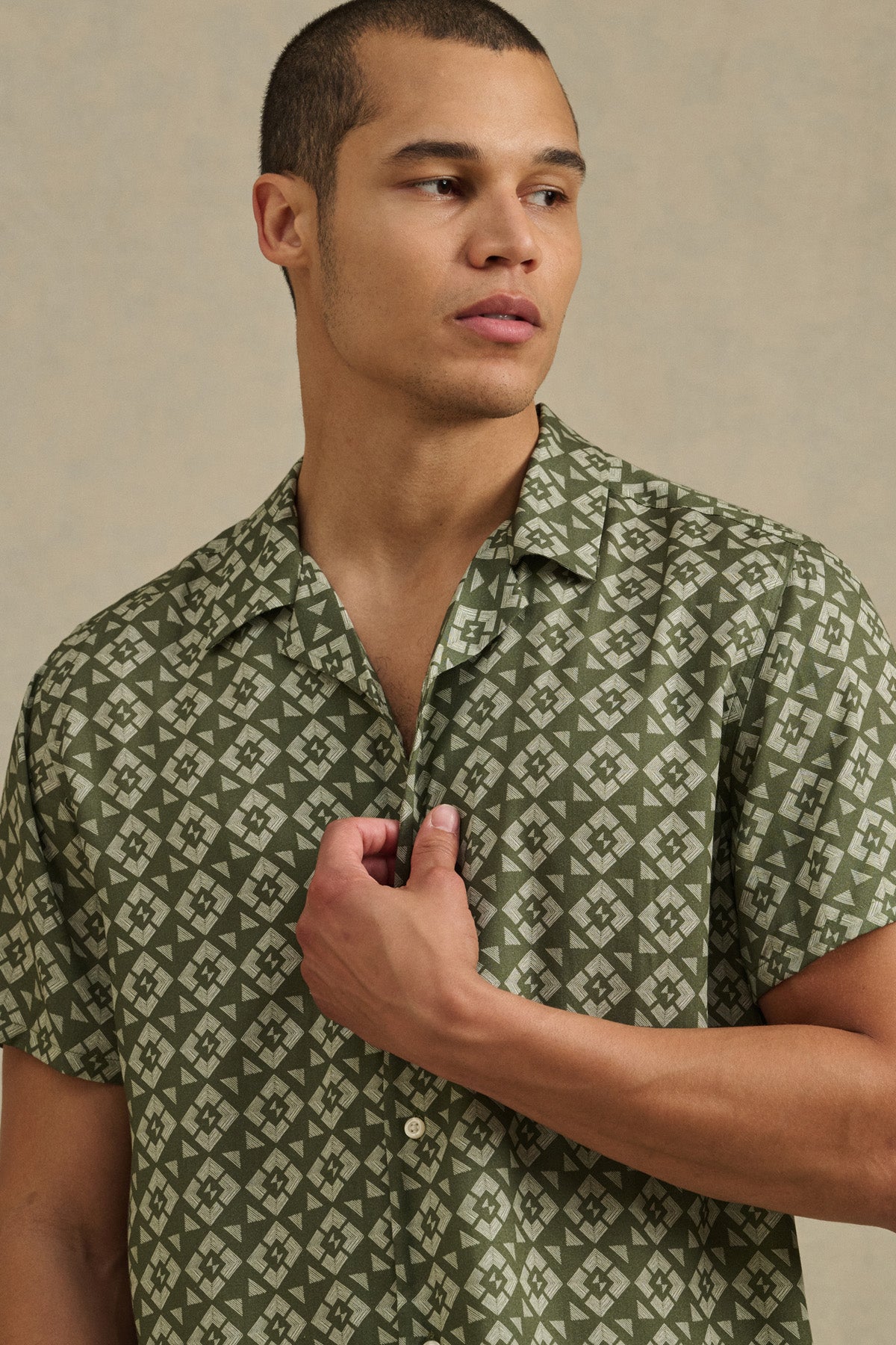 A man wearing the EMILIO SHIRT by Velvet by Graham & Spencer—a short-sleeved, green camp collar shirt with a relaxed fit—stands against a plain background, looking left with one hand touching his shirt.-41479916421313