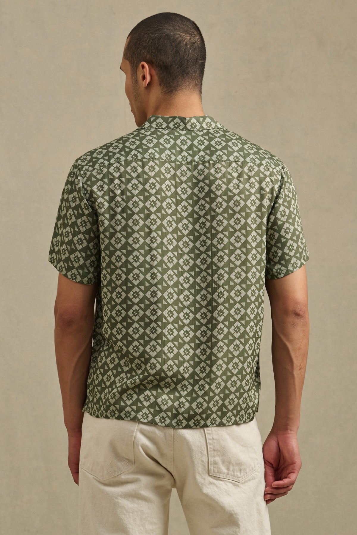 A person stands facing away in the Velvet by Graham & Spencer EMILIO SHIRT—a green camp collar shirt with a geometric white pattern—paired with light-colored pants against a neutral background.