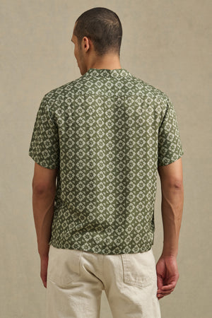 A person stands facing away in the Velvet by Graham & Spencer EMILIO SHIRT—a green camp collar shirt with a geometric white pattern—paired with light-colored pants against a neutral background.