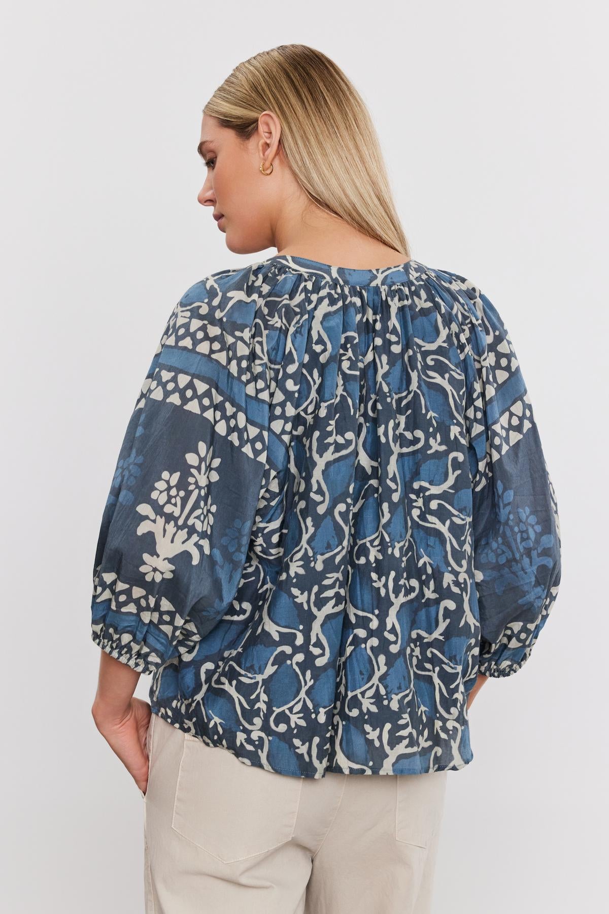 Woman from behind wearing a blue patterned Dayana top by Velvet by Graham & Spencer with elastic cuffs and puffed sleeves, paired with khaki pants, standing against a white background.-36918801465537