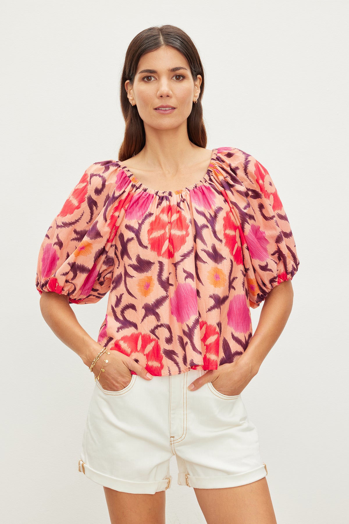   Woman standing against a plain background wearing the EDLIN PRINTED SILK COTTON VOILE TOP by Velvet by Graham & Spencer with puff sleeves and white shorts. Crafted from printed silk cotton, the blouse exemplifies versatile styling. She has long, straight hair and is looking directly at the camera. 