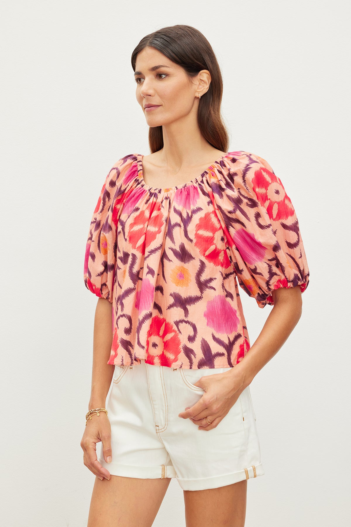   A woman wearing a vibrant, floral-patterned Velvet by Graham & Spencer EDLIN PRINTED SILK COTTON VOILE TOP with puff sleeves and white shorts stands against a plain background. The printed silk cotton fabric adds a touch of elegance as she gazes to her left. 