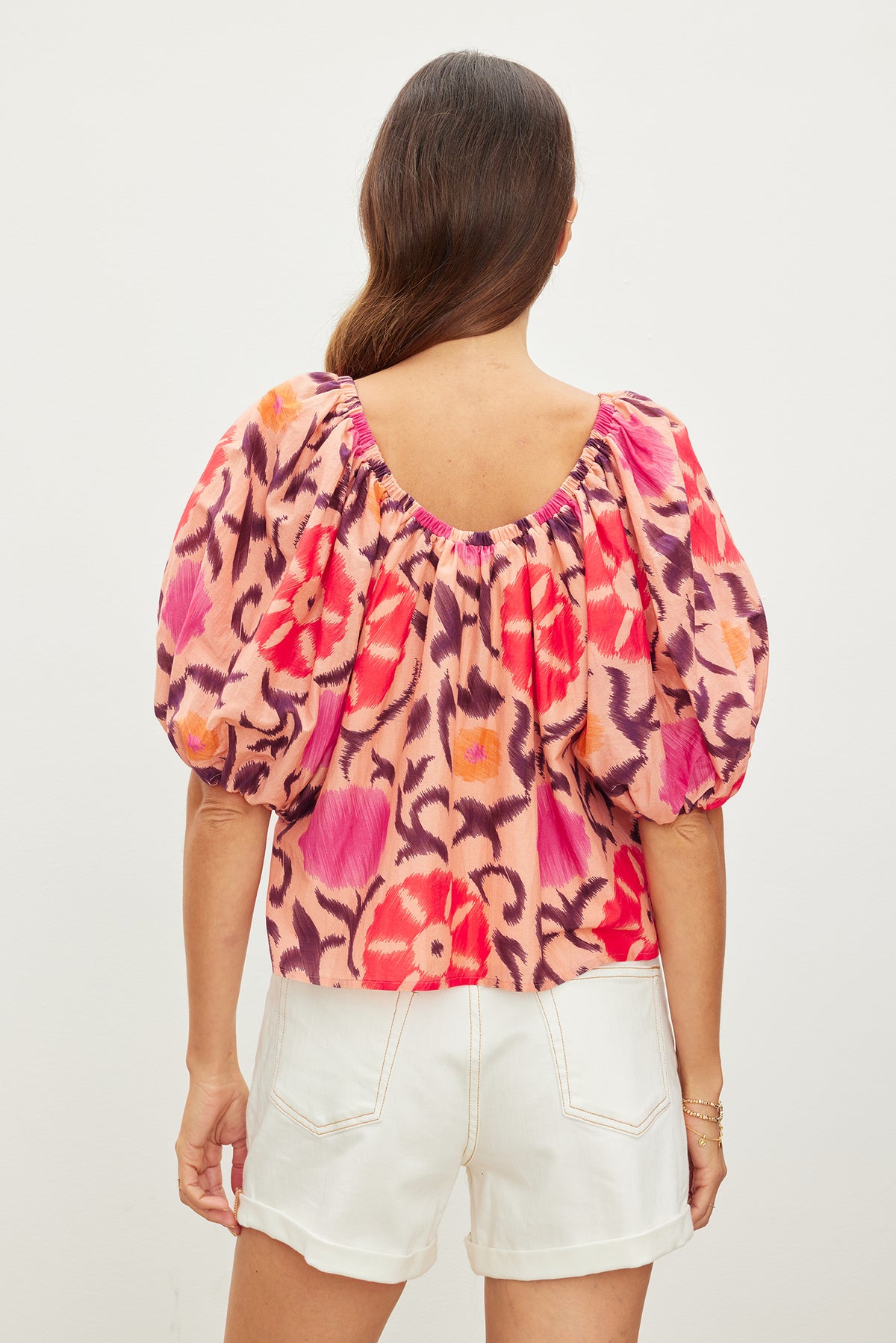   Woman in white shorts and a colorful EDLIN PRINTED SILK COTTON VOILE TOP by Velvet by Graham & Spencer stands facing away, showcasing the blouse's back design. 