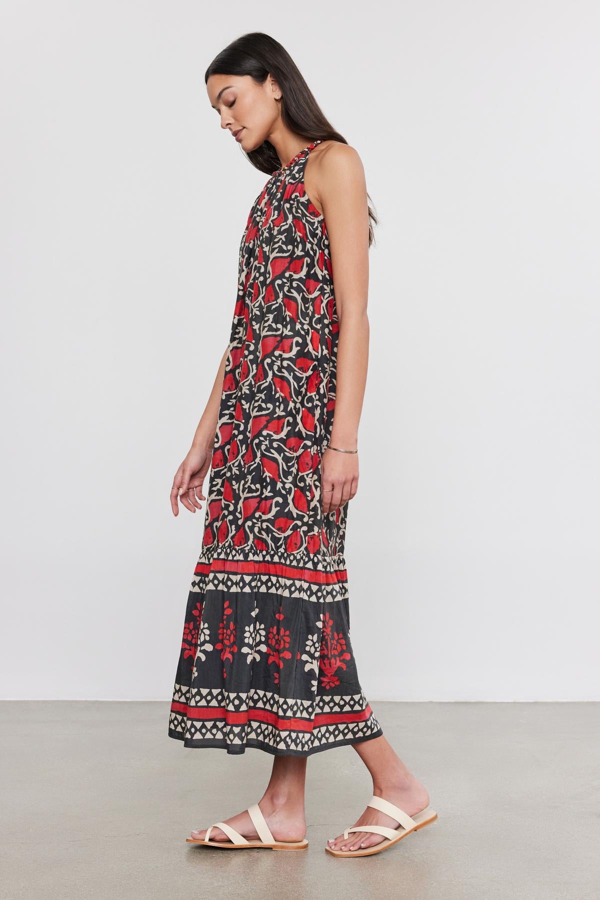   A woman wearing a sleeveless, ankle-length GHITA DRESS by Velvet by Graham & Spencer with a red, black, and white ethnic print, paired with white sandals, stands looking down. 