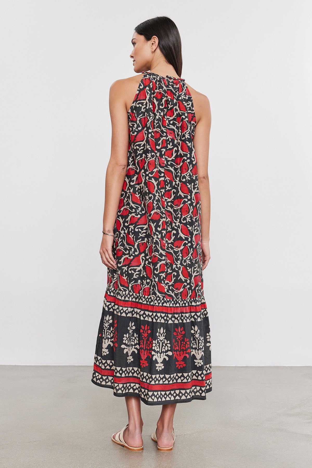   Woman standing, viewed from the back, wearing a long, sleeveless black silk cotton voile GHITA DRESS by Velvet by Graham & Spencer with bold red and white floral prints, standing in a plain studio setting. 