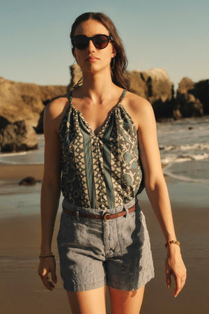 A woman in sunglasses, wearing a Velvet by Graham & Spencer RHEA TANK TOP with a v-neckline and denim shorts, stands on a sandy beach with cliffs in the background.