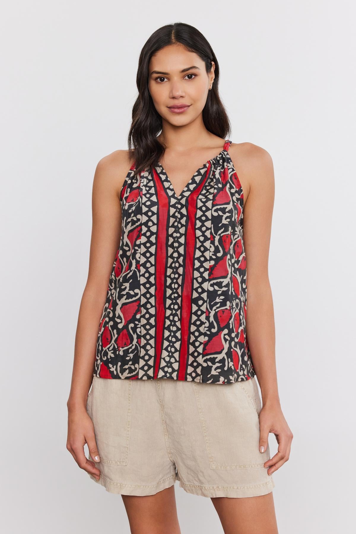   A woman wearing the RHEA TANK TOP by Velvet by Graham & Spencer, designed with a red, black, and beige pattern and paired with beige shorts. The printed silk cotton voile top features a v-neckline that creates a flattering silhouette. She stands against a plain white background. 