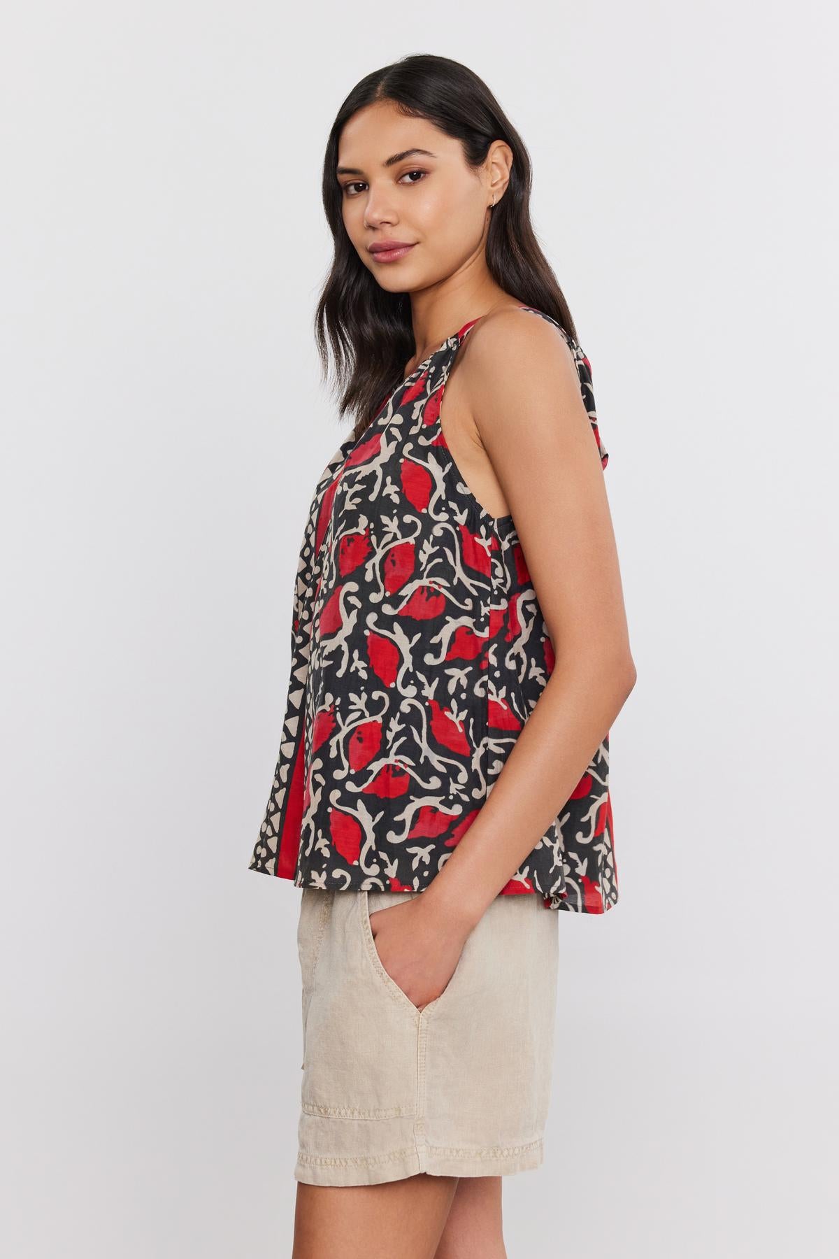   A woman with long dark hair stands sideways, wearing a RHEA TANK TOP by Velvet by Graham & Spencer, a sleeveless black, red, and white patterned top that flatters her silhouette, paired with light beige shorts. Her hands are in her pockets against a plain white background. 