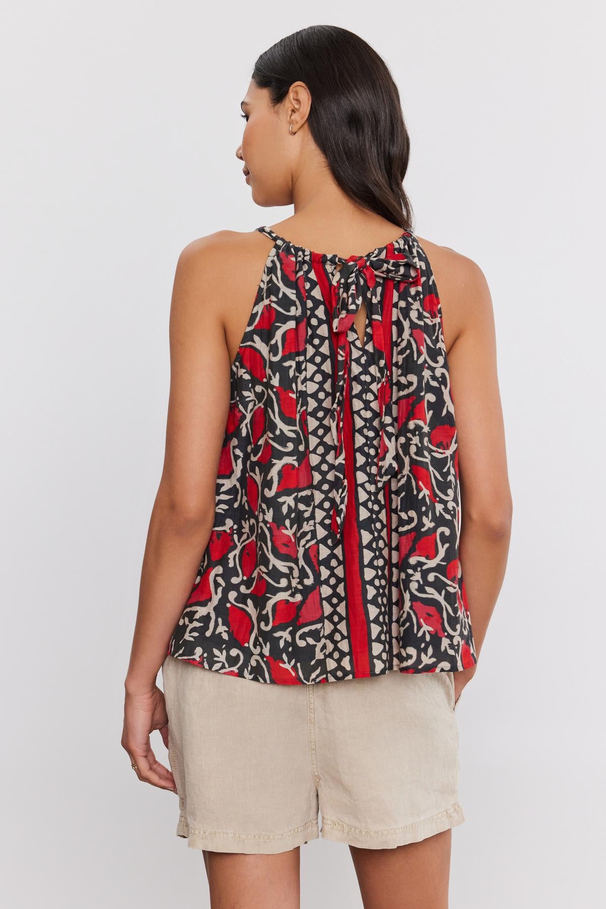   A woman with dark hair is seen from the back, wearing a sleeveless RHEA TANK TOP from Velvet by Graham & Spencer, featuring a patterned design and a tie at the neck. She pairs it with beige shorts. The v-neckline of her tank top contributes to its flattering silhouette, enhancing her elegant appearance. 