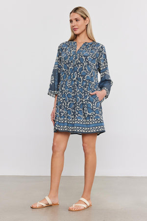 A woman models a blue printed tiered Talia dress with 3/4 sleeves and white sandals, standing against a plain white background from Velvet by Graham & Spencer.