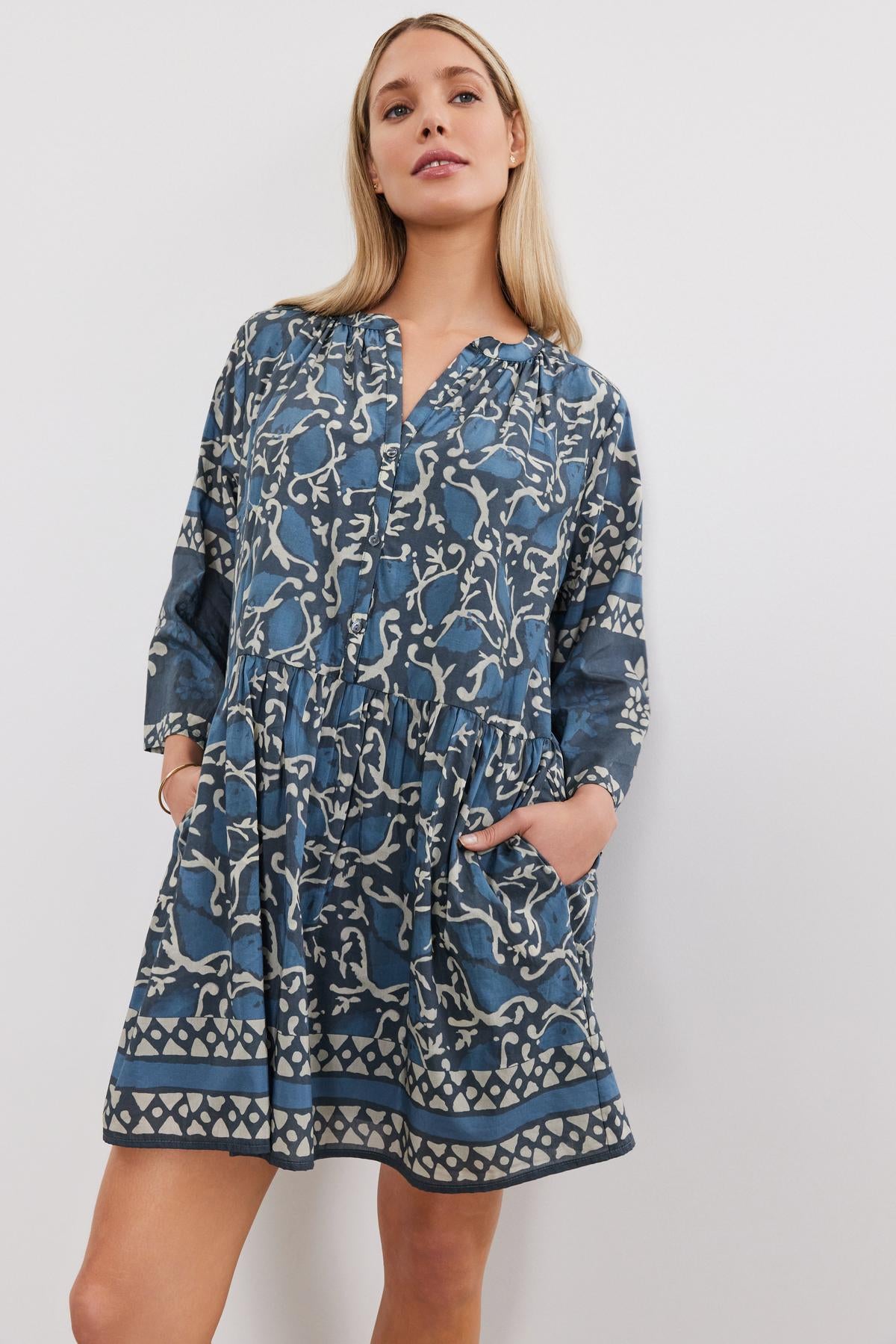   A woman wearing a blue and white patterned split neck Talia dress with long sleeves, standing against a white background by Velvet by Graham & Spencer. 