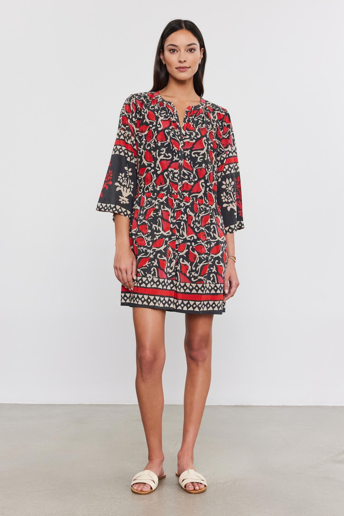   A person stands against a plain background, wearing the TALIA DRESS by Velvet by Graham & Spencer—a black and red patterned split neck dress with bell sleeves—paired with white sandals. 