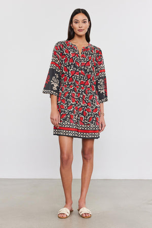 A person stands against a plain background, wearing the TALIA DRESS by Velvet by Graham & Spencer—a black and red patterned split neck dress with bell sleeves—paired with white sandals.