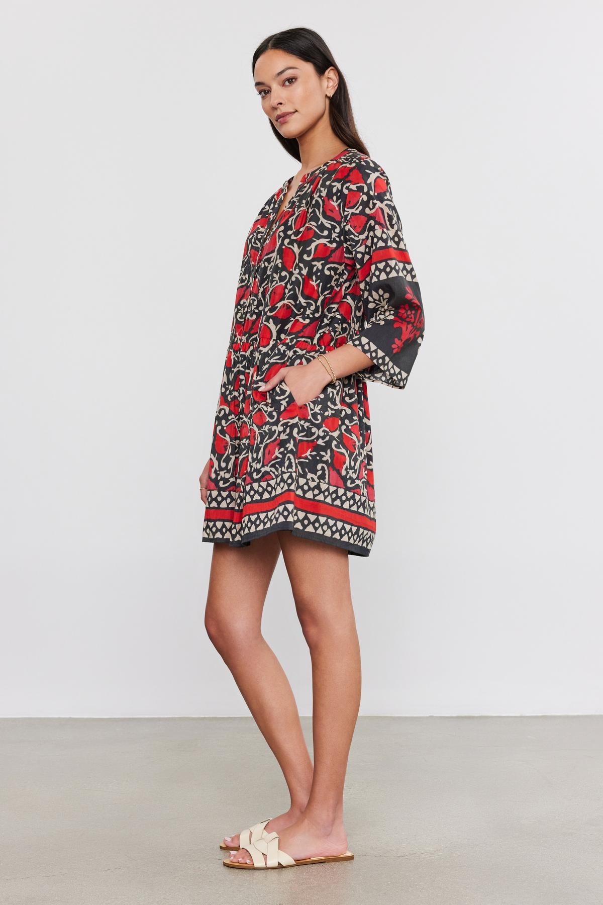   A woman stands in a relaxed pose wearing the TALIA DRESS by Velvet by Graham & Spencer. The knee-length dress features a split neck and red, black, and white floral patterns on printed silk cotton voile fabric that flows gracefully as she rests her hands in the pockets. She completes the look with beige flat sandals. 