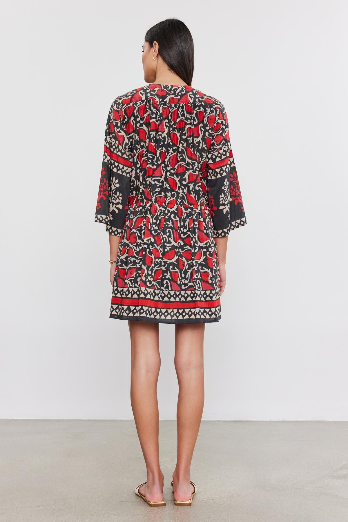   Back view of a person with long dark hair wearing the TALIA DRESS from Velvet by Graham & Spencer, a floral-patterned silk cotton voile dress with red and black tones and 3/4 sleeves, standing against a plain white background. 