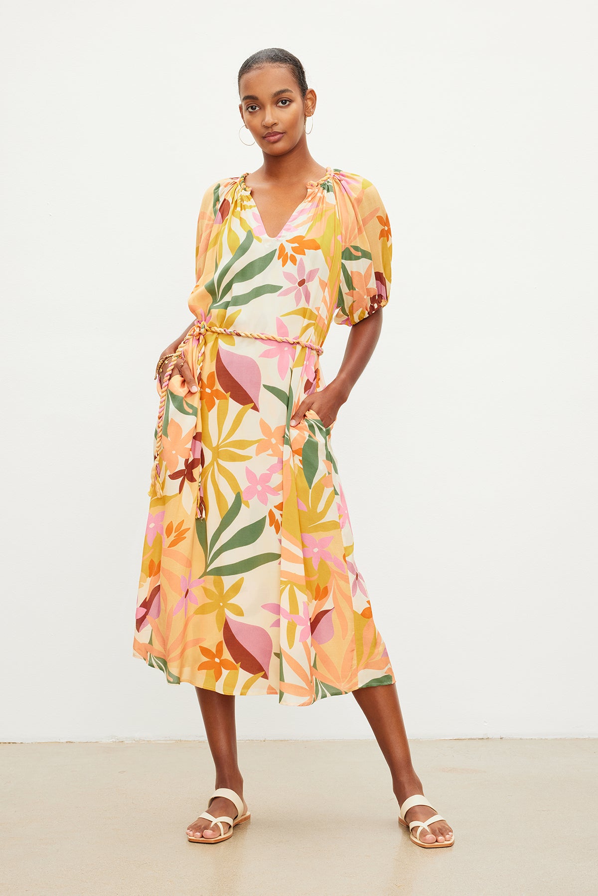   A woman standing, wearing a colorful floral printed CAROL BOHO DRESS by Velvet by Graham & Spencer and beige sandals, with her hands gently resting on her hips. 