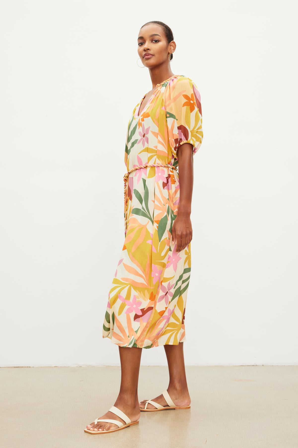 A person wearing the CAROL PRINTED BOHO DRESS by Velvet by Graham & Spencer, featuring a brightly patterned, short-sleeve viscose design with a v-neckline and paired with sandals, is standing on a plain background and viewed from a side angle.-37618694324417