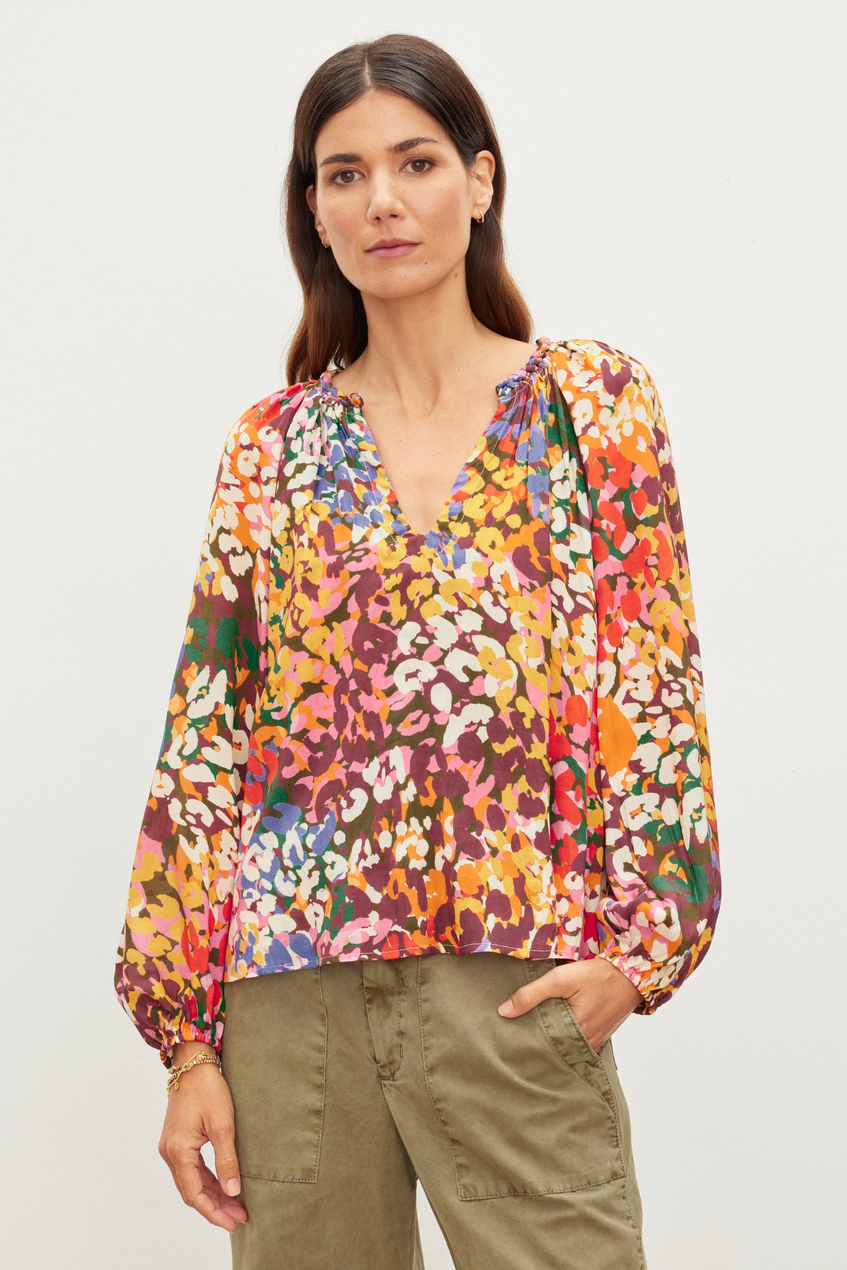   A person wearing a colorful floral DION TOP by Velvet by Graham & Spencer with a v-neckline and long sleeves featuring elastic cuffs, paired with relaxed fit olive green pants, stands against a plain white background. 