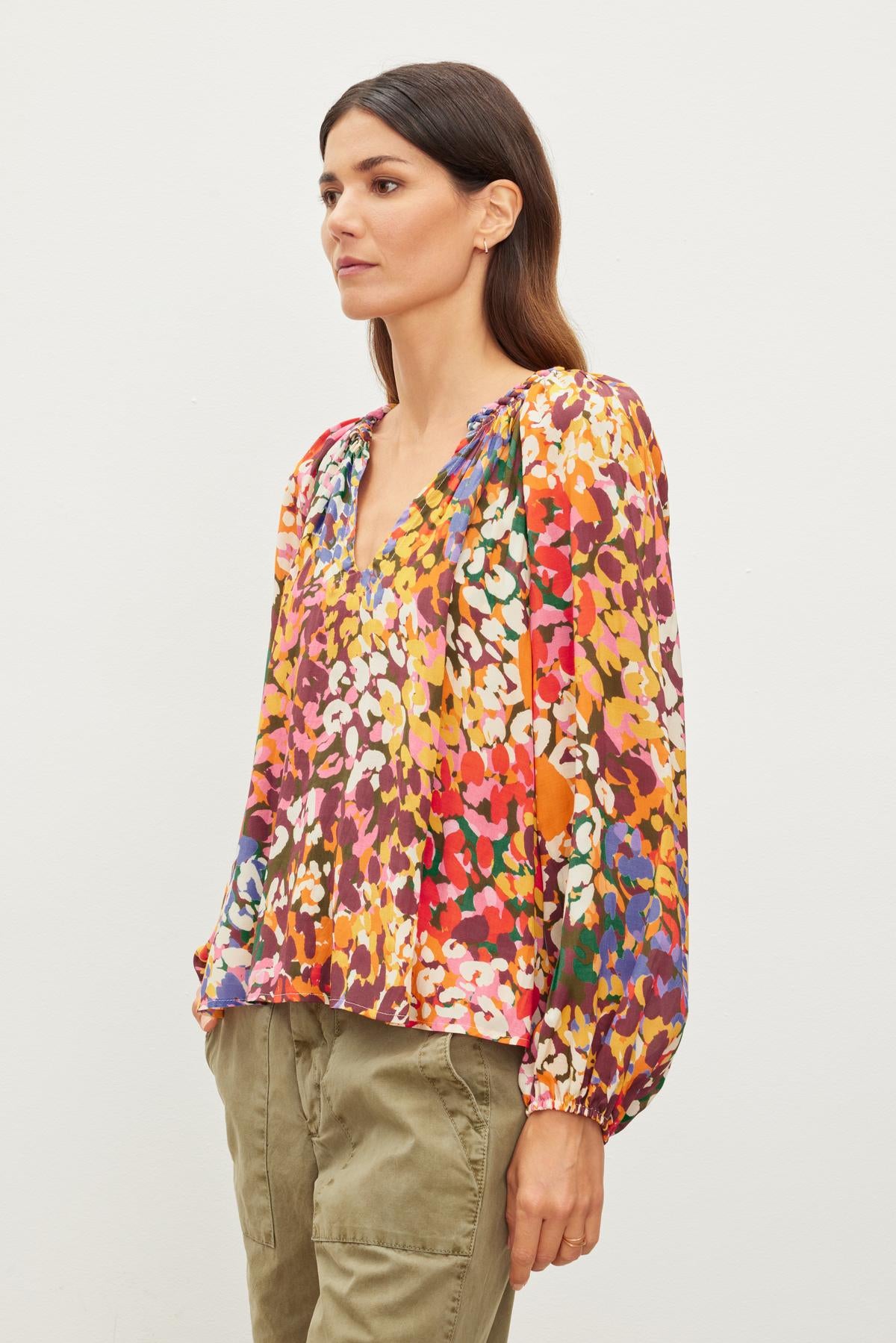   A person with long dark hair wearing the DION TOP by Velvet by Graham & Spencer, which is a colorful, patterned blouse with elastic cuffs, and olive green pants in a relaxed fit stands against a plain white background. 