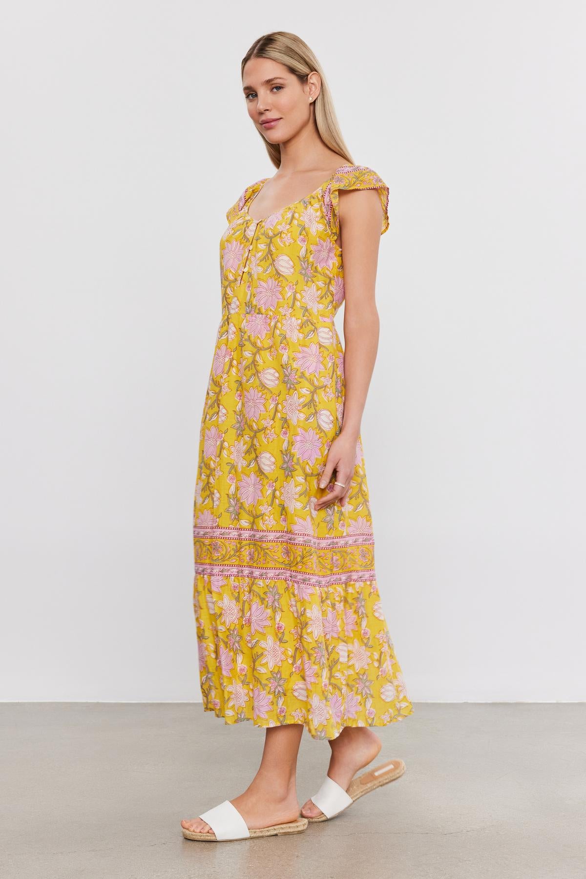   A woman in the JAZLYN DRESS by Velvet by Graham & Spencer, a midi-length yellow floral-printed cotton voile dress, paired with white sandals, stands against a plain background. 