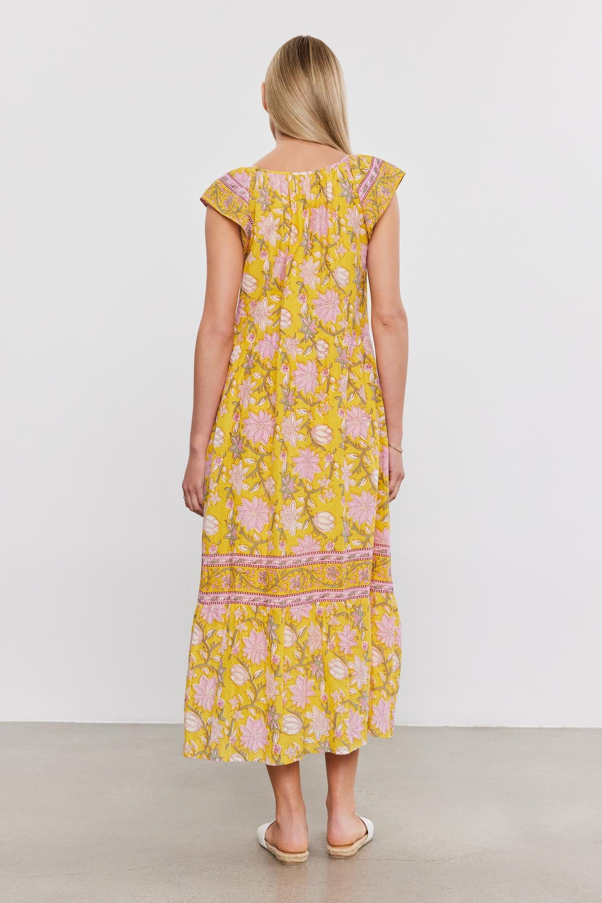   A woman with blonde hair stands facing away, showcasing the back of her JAZLYN DRESS by Velvet by Graham & Spencer. The yellow floral midi-length dress features charming cap sleeves, and she accessorizes it with white sandals. She stands on a plain gray floor against a white wall background, capturing the essence of a perfect summer wardrobe. 