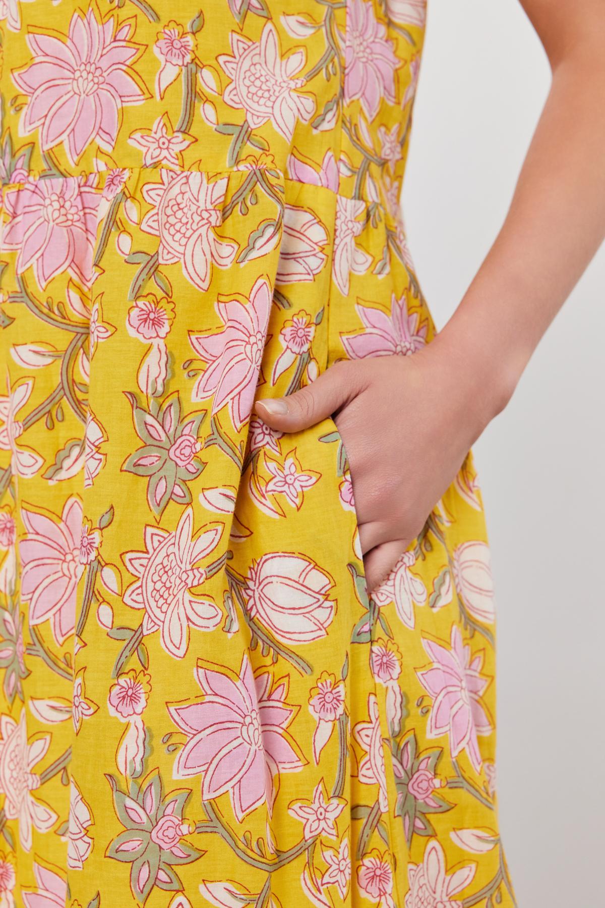   Close-up of a person wearing the JAZLYN DRESS by Velvet by Graham & Spencer, showcasing its yellow printed cotton voile fabric adorned with pink floral patterns. The right hand rests in the pocket of this stylish midi-length dress—perfect for any summer wardrobe. 