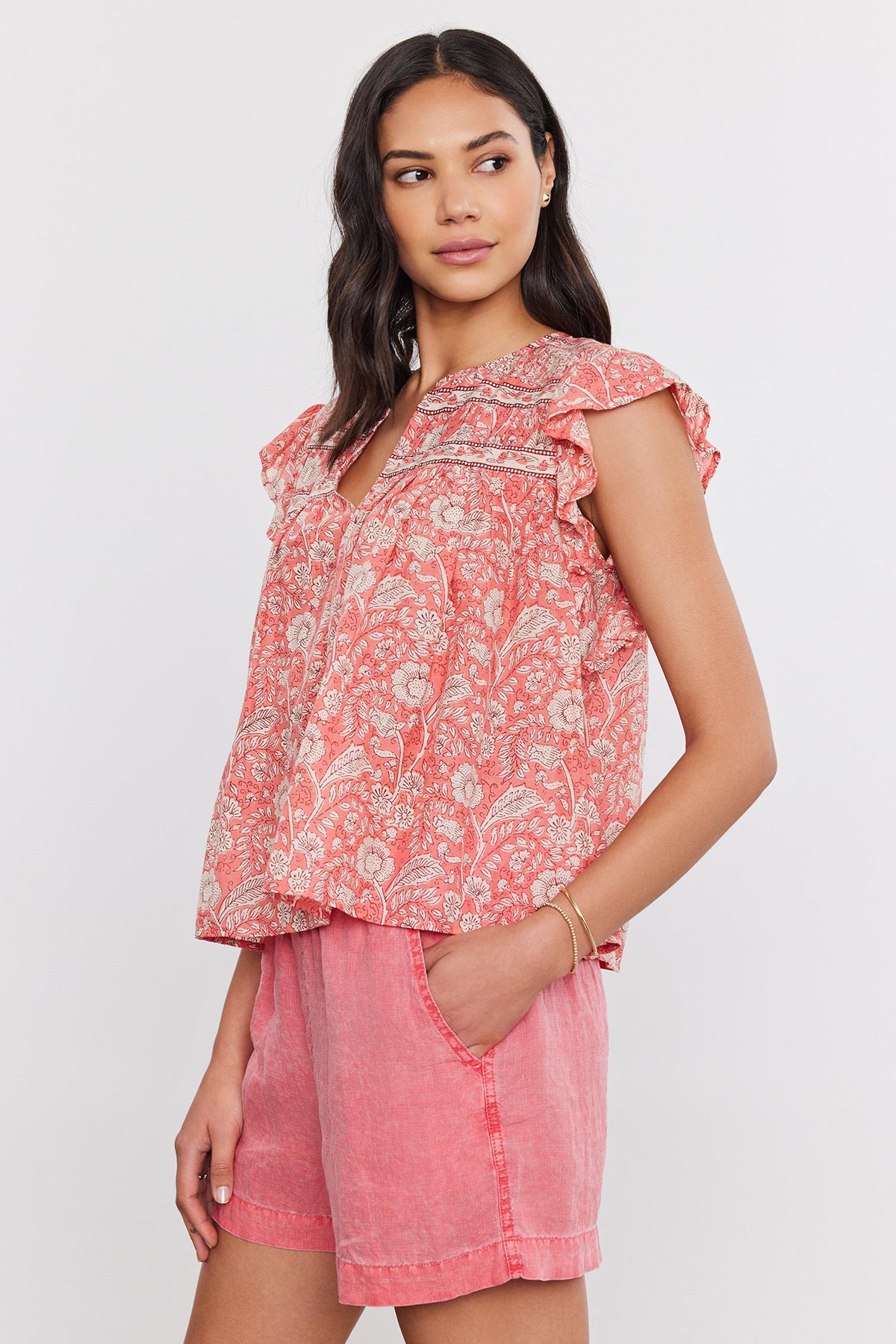 Woman in a pink floral print Kenzie top by Velvet by Graham & Spencer and denim shorts, standing with one hand on her hip, looking towards the camera.-36910087405761