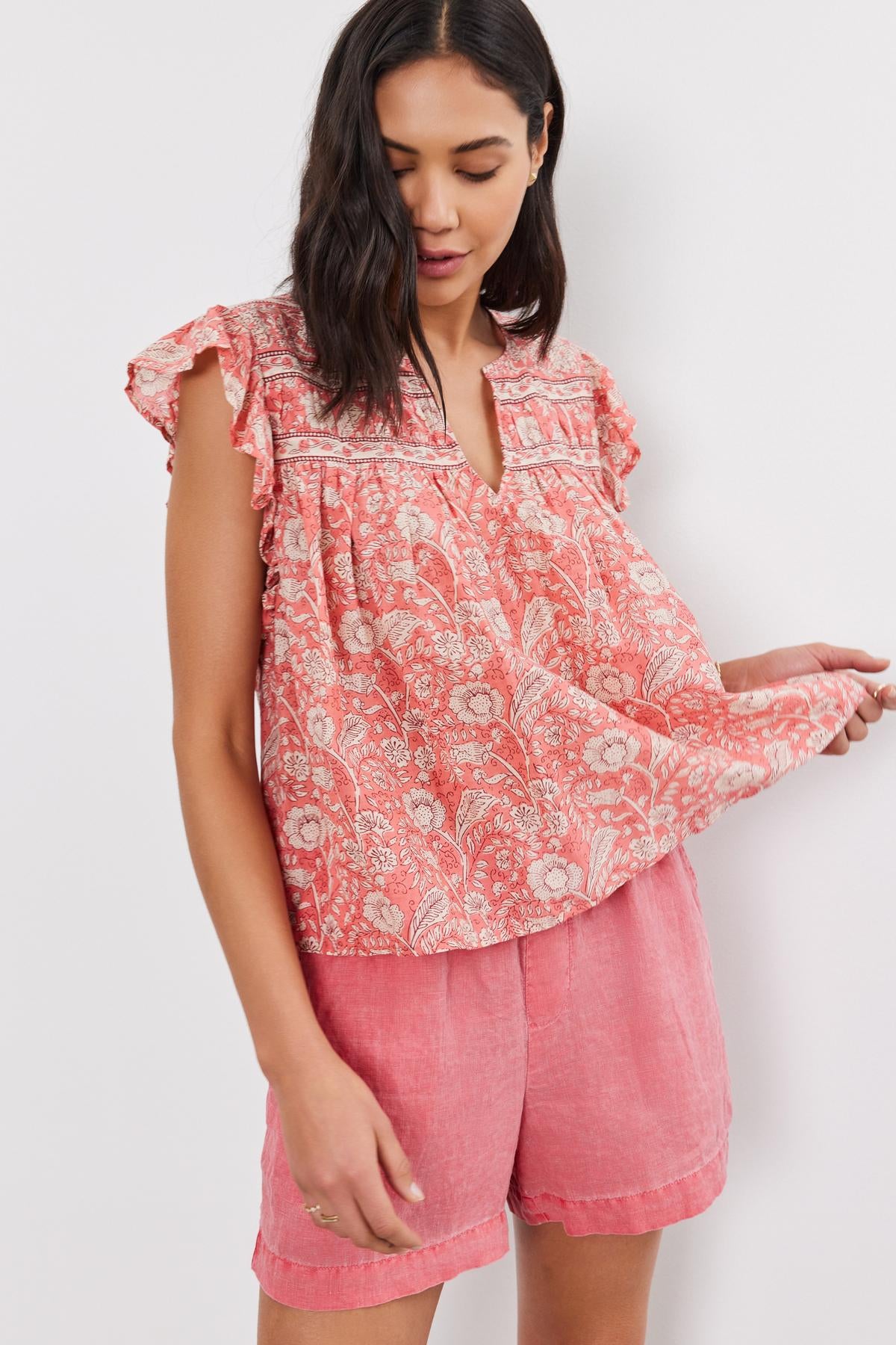 A woman wearing a pink Velvet by Graham & Spencer Kenzie Top with floral print and matching shorts, standing against a white background, examining her sleeve.-36910087504065