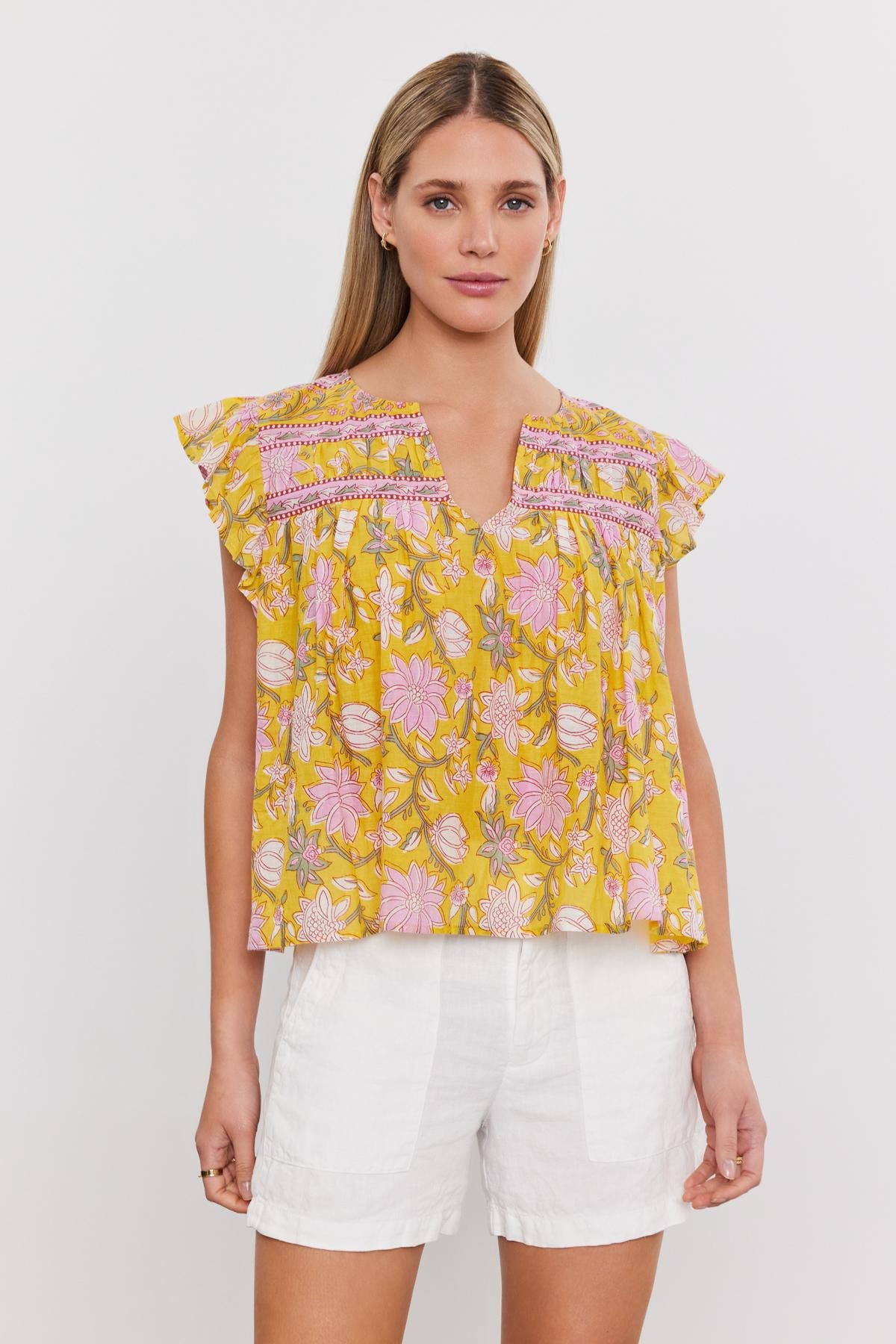   A woman is standing against a plain white background, wearing the KENZIE TOP by Velvet by Graham & Spencer, which is yellow with pink accents and features a delightful floral print, paired with white shorts. 