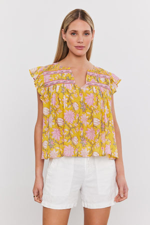 A woman is standing against a plain white background, wearing the KENZIE TOP by Velvet by Graham & Spencer, which is yellow with pink accents and features a delightful floral print, paired with white shorts.