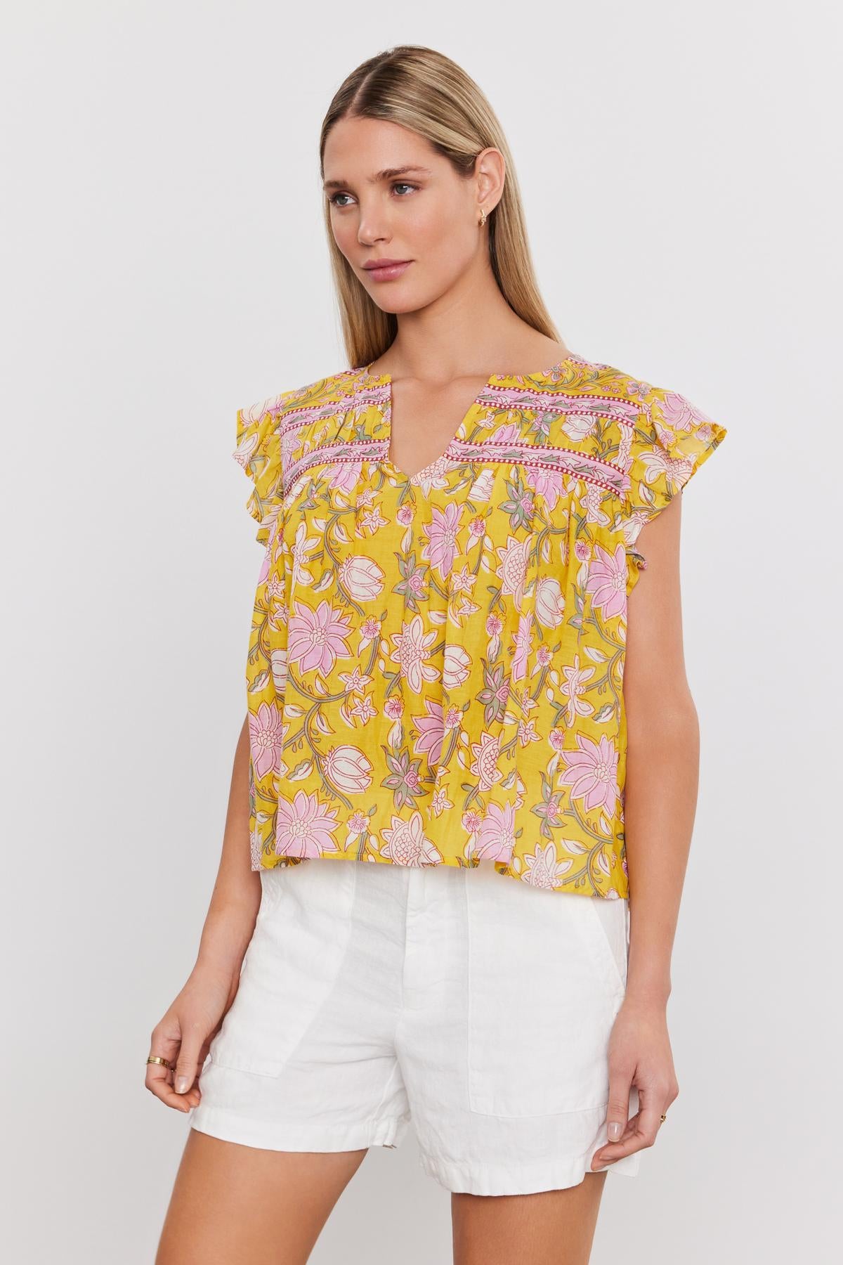   A woman is wearing a vibrant yellow floral KENZIE TOP from Velvet by Graham & Spencer, featuring charming ruffled sleeves, paired with stylish white shorts—ideal for a summer outfit. She has straight, long blonde hair and is posed against a plain white background. 