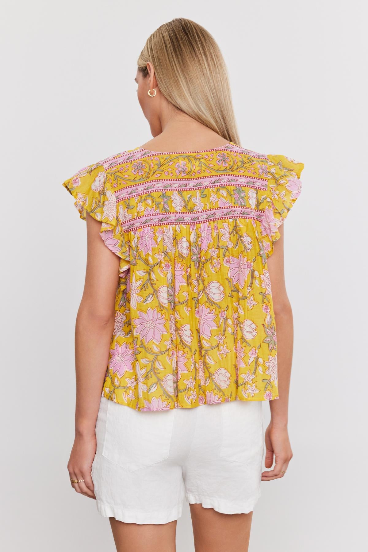   A person is shown from the back wearing the KENZIE TOP by Velvet by Graham & Spencer, featuring a yellow and pink floral print with ruffled sleeves, paired with white shorts. The person's hair is straight and worn down, creating a perfect summer ensemble. 