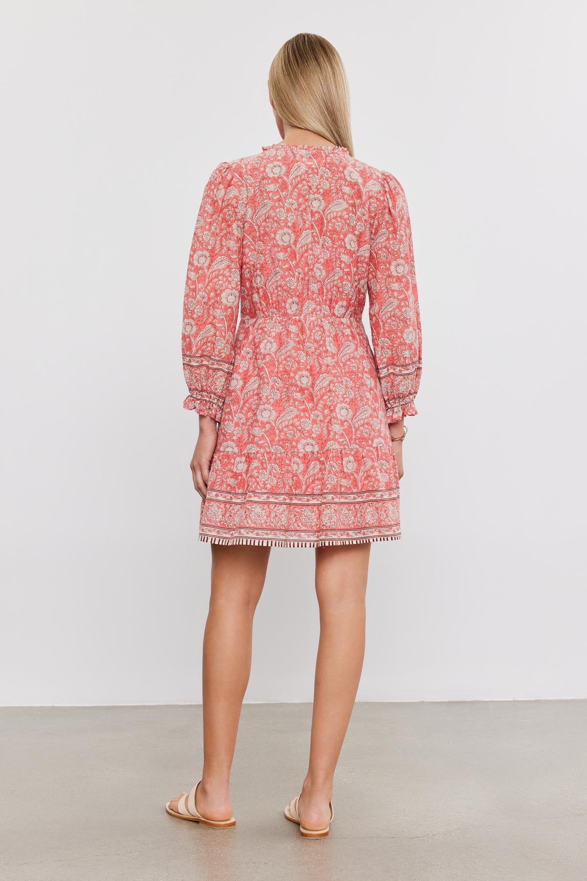   A woman stands facing away from the camera, wearing a pink floral tiered MARY DRESS with long sleeves and fringed hem, paired with light beige flats from Velvet by Graham & Spencer. 