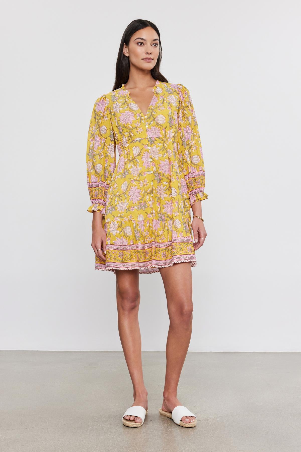   A woman stands against a plain background, wearing the MARY DRESS from Velvet by Graham & Spencer. The vibrant printed cotton voile dress features long sleeves, and she pairs it with white sandals. Facing forward, her left foot is slightly ahead of the other. 