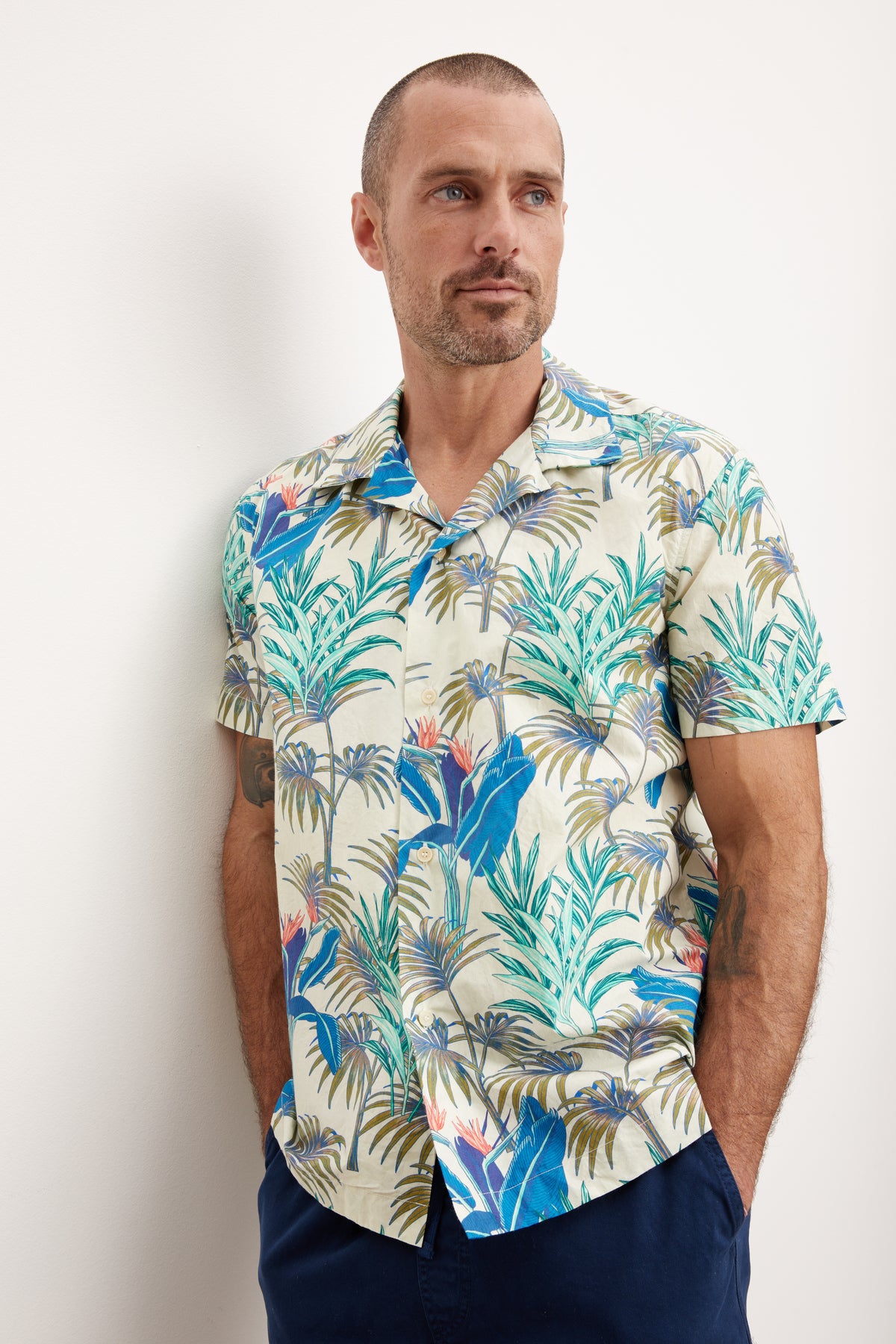   A man in a Velvet by Graham & Spencer IGGY BUTTON-UP SHIRT stands against a white wall, looking to the side with a thoughtful expression. 