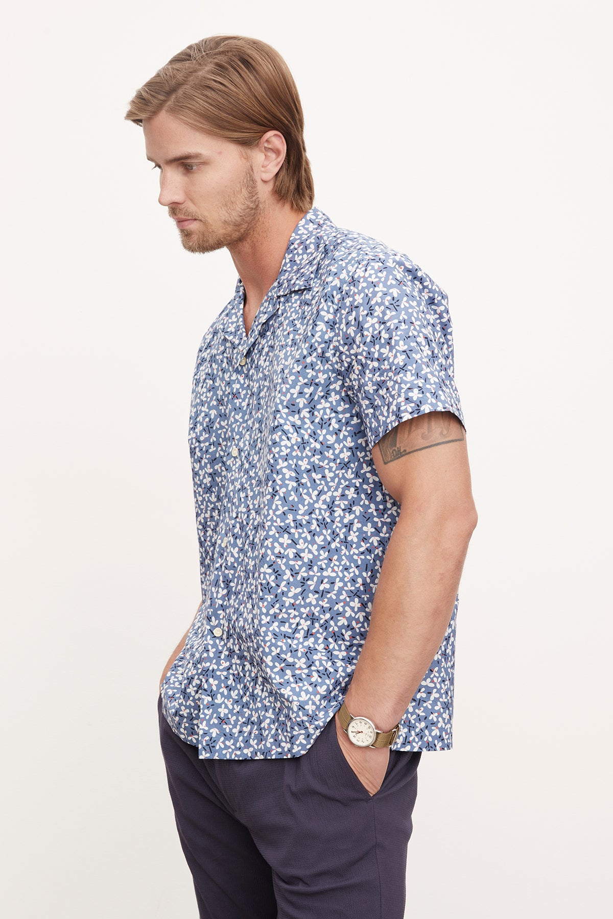   Man in a blue and white floral print Iggy button-up shirt by Velvet by Graham & Spencer and navy trousers, standing sideways, looking to the left with a watch on his wrist. 
