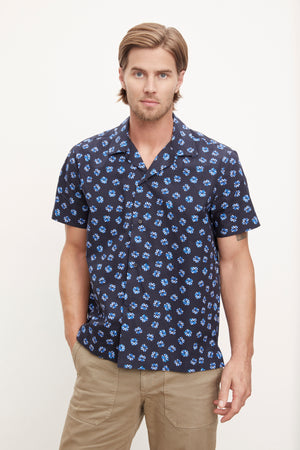 Man wearing a short-sleeved patterned Iggy button-up shirt by Velvet by Graham & Spencer and khaki pants standing against a plain background.