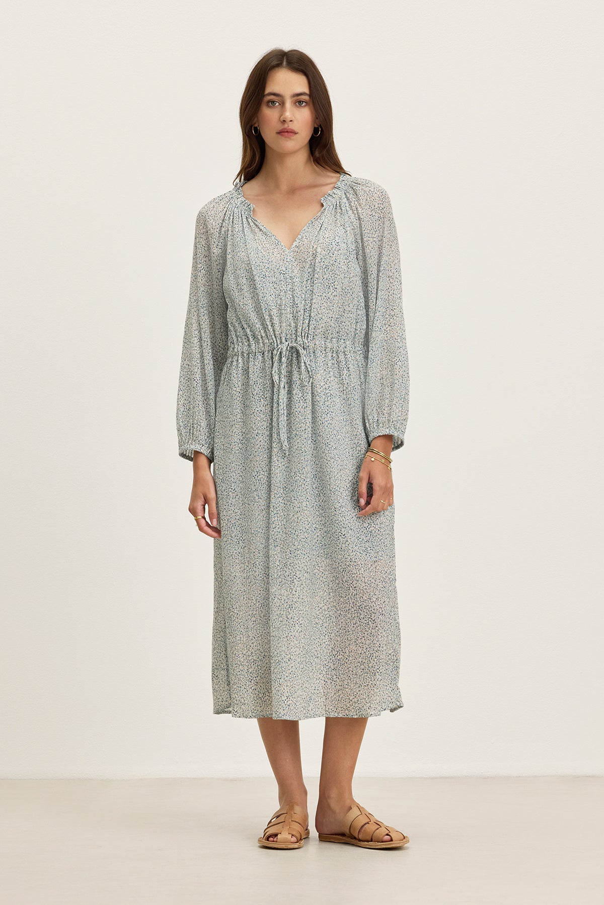 A woman stands against a plain background wearing the LIMA PRINTED CRINKLE COTTON GAUZE DRESS by Velvet by Graham & Spencer, featuring a ruffled V-neckline, drawstring waist, and tan sandals.-39333213569217