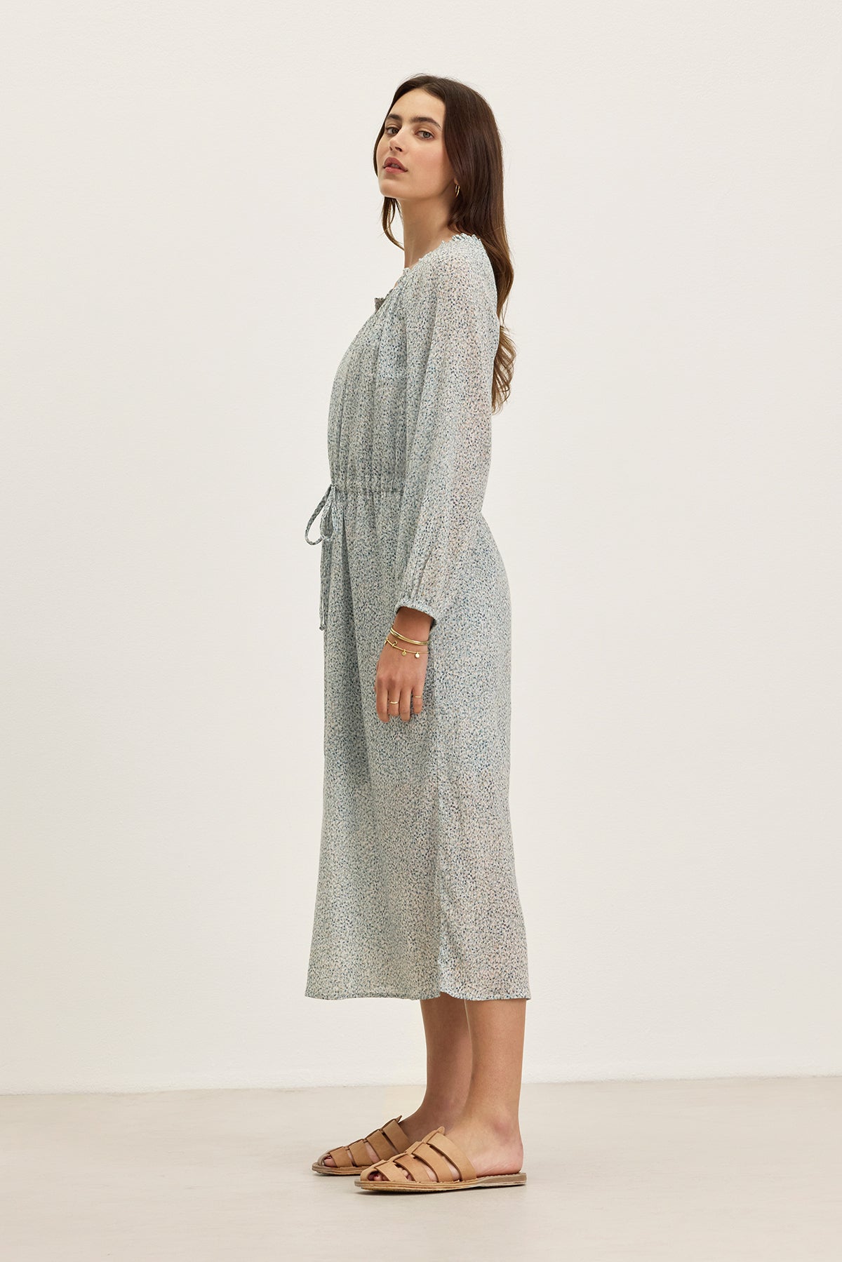 A woman stands sideways wearing the LIMA PRINTED CRINKLE COTTON GAUZE DRESS by Velvet by Graham & Spencer, featuring a light blue print, ruffled V-neckline, waist tie, and tan flat sandals against a plain background.-39333213601985