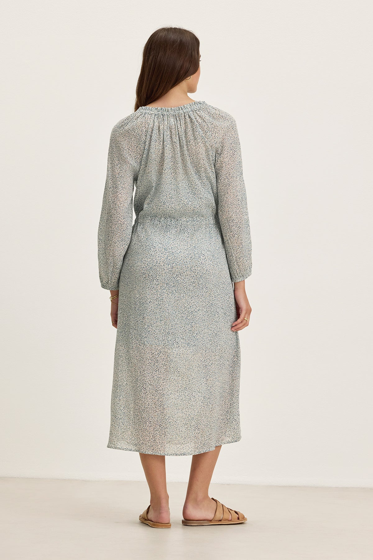 A woman faces away in the Velvet by Graham & Spencer LIMA PRINTED CRINKLE COTTON GAUZE DRESS—a light blue, patterned midi with a ruffled V-neckline—paired with tan flat sandals against a plain light background.-39333213634753